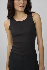 Frau trägt ein enganliegendes, schwarzes, ärmelloses, geripptes Oberteil mit einem dezenten, gestickten Logo auf der Brust und maßgeschneiderte schwarze Hosen.