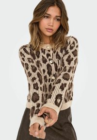 Mlada žena nosi kardigan sa leopard uzorkom u bež i smeđim tonovima s velikim dugmadima, i tamnu suknju, pozira s prekriženim rukama ispred sebe.