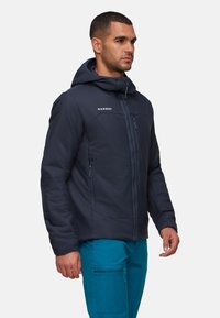 Uomo che indossa una giacca con cappuccio blu navy con tasche con zip e pantaloni turchesi, in piedi contro uno sfondo chiaro e uniforme.