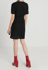 Schwarzes Kurzarmkleid mit taillierter Passform und einem dezenten Schimmer, kombiniert mit glänzenden roten Knöchelstiefeln mit einem klobigen Absatz.