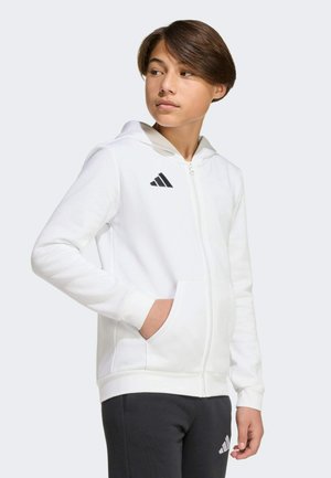 Młoda osoba nosząca białą bluzę z kapturem na zamek z czarnym logo Adidas, jedną ręką w kieszeni, patrząca w bok, ubrana w czarne spodnie.