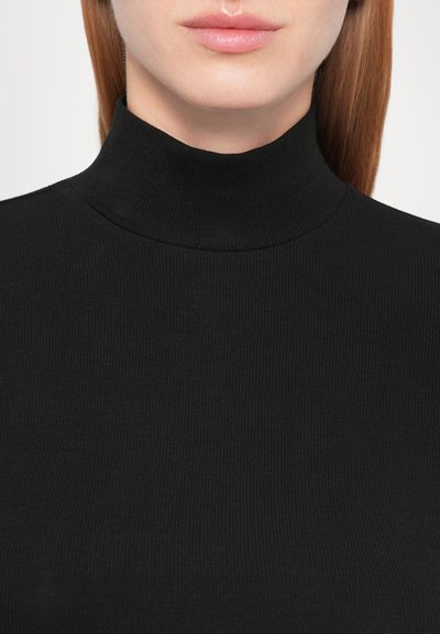 Gros plan d'une personne portant un pull noir côtelé à col roulé, montrant les lèvres, le menton et le cou avec des cheveux bruns raides.