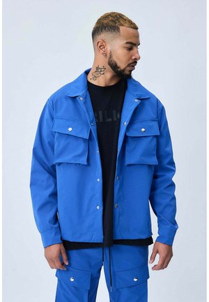 Homme aux cheveux teints et tatouage au cou, portant une veste bleu vif avec de grandes poches et un pantalon assorti, regardant sur le côté.