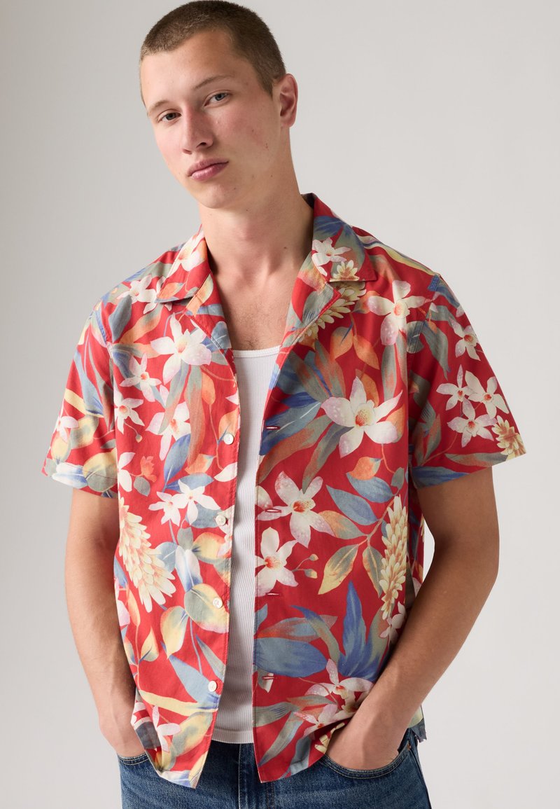 Joven con pelo corto que lleva una camisa roja de flores de manga corta sobre una camiseta blanca sin mangas, manos en los bolsillos, sobre un fondo liso.