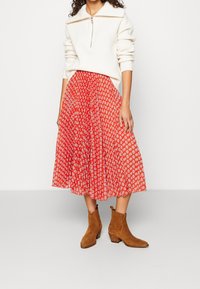 Roter, plissierter Rock mit weißem Blumenmuster, kombiniert mit einem cremefarbenen Pullover mit Reißverschluss am Ausschnitt. Braune Stiefeletten mit Absatz.