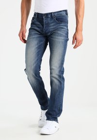 Jeans de denim azul con un corte slim, acabado desgastado y detalles de costura. Presenta cinco bolsillos y una cremallera con un botón de metal.