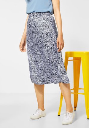 Vrouw die een midi-rok met een blauw-wit bladpatroon draagt, een lichtblauw topje en witte sneakers, staand naast een gele metalen kruk.