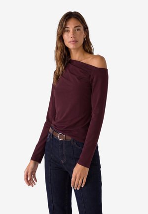 Kvinde iført en bordeauxfarvet asymmetrisk langærmet top og mørkeblå jeans med brunt bælte, stående mod en hvid baggrund.