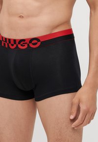 Black boxer shorts with a red elastic waistband featuring the logo "HUGO." The fabric appears smooth with a snug fit.