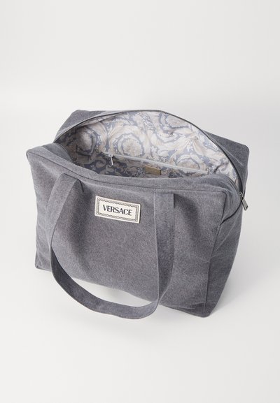 Saco tote de tecido cinza com duas alças e fecho em zíper. O interior apresenta um padrão floral azul claro. Logo da Versace na frente.