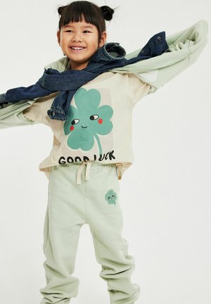 Lächendes Kind mit Zöpfen, trägt einen beigen Sweatshirt und eine Hose mit kleinem lächelnden vierblättrigen Kleeblatt und dem Text "Viel Glück", hält eine Jeansjacke in der Hand.