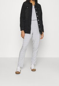 Černá džínová bunda s předními knoflíky, kombinovaná s bílým overalem s vzorem. Bílá teniska s hnědými podrážkami doplňuje outfit.