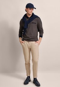 Dunkelgraues Langarmshirt mit marineblauen Akzenten, beigen maßgeschneiderten Hosen, marineblauem Hut und Schuhen. Ein marineblauer Pullover ist um den Hals drapiert.