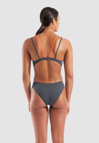 Femme aux cheveux bruns attachés portant un bikini gris foncé avec de fines bretelles orange et grises, se tenant devant un fond gris clair uni.
