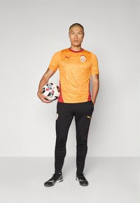 Oranssi-punainen Puma urheilupaita, jossa on tekstuuri kuvio, mustat Puma-housut punaisilla sivuraitoilla ja mustavalkoinen jalkapallo.