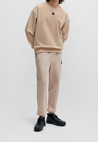 Sudadera beige con escote redondo y un pequeño parche con el logo, combinada con pantalones de chándal beige a juego. Zapatillas negras completan el atuendo.