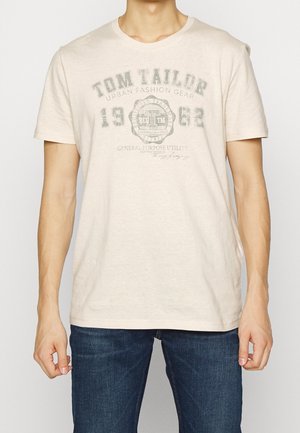 Homme portant un t-shirt beige Tom Tailor à manches courtes avec un texte imprimé et un jean bleu foncé, les bras détendus le long du corps.