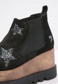 Botas de tobillo de ante negro con paneles laterales de elasticidad negra, patrones de estrellas brillantes, un tacón de cuña texturizado en bronce y suela de goma negra con bordes irregulares.