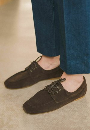 Scarpe in suede marrone con profilo basso, punta rotonda e chiusura con lacci. Le scarpe presentano una tomaia testurizzata e una suola in gomma beige.