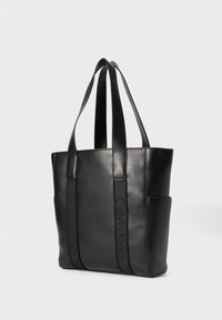 Sac fourre-tout en cuir noir avec deux longues poignées, présentant des accents cousus et une poche latérale. Affiche le logo Calvin Klein dans un design tonal.