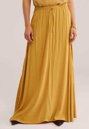 Žena má na sebe maxi sukňu horčicovej farby so šnúrkou v páse a hnedé špicaté topánky, stojí s uvoľnenými rukami pri bokoch.