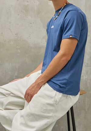 Person, die ein blaues T-Shirt und weiße Hose trägt, sitzt auf einem Holzschemel vor einer grauen Betonwand.