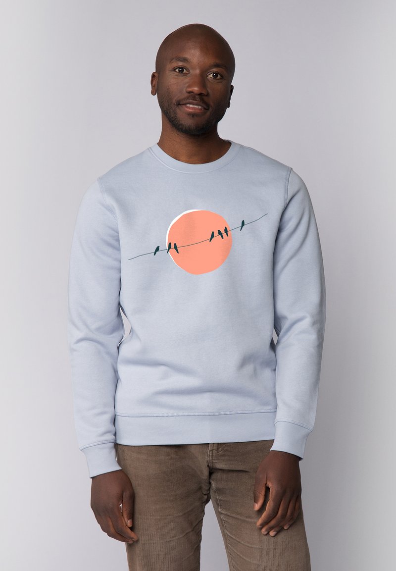 Hellblaues Sweatshirt mit einem runden, orangen Grafik, die einen Sonnenuntergang und Vögel auf einer Leitung darstellt. Weicher Stoff, lässige Passform, gerippte Bündchen und Saum.