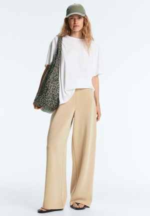 Žena oblečená v bielej veľkej tričku, béžových širokých nohaviciach, čiernych sandáloch, zelenej čiapke a nesie veľkú tašku s leopardím vzorom.