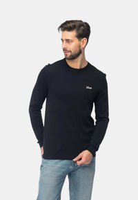 Maglione nero a maniche lunghe con scollo rotondo, realizzato in morbido tessuto, con un piccolo logo sul petto e polsini a costine, abbinato a jeans blu chiaro.