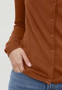 Brun knapplistad cardigan med ribbade manschetter, mjuk textur och en figurnära design, bärs över blå denim. Knapparna är runda och matchande i färg.