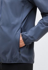 Άτομο που φοράει μπλε μπουφάν με ελαστικά μανσέτα, το χέρι μέσα σε τσέπη με φερμουάρ στο πλάι, πάνω σε απλό φωτεινό φόντο.