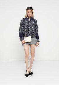 Woman in floral navy blouse and gray mini skirt holding white clutch, wearing black pointed heels with bows, standing against white background.