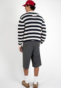 Hombre con gorra roja, suéter de punto a rayas azul marino y blanco, pantalones cortos grises con rayas finas hasta la rodilla, calcetines blancos y zapatos marrones de espaldas.