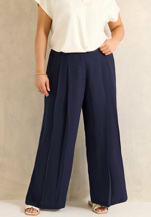 Vrouw die wijde marineblauwe broek draagt, witte korte mouwen blouse, witte open-toe sandalen en een gouden armband, staande tegen een effen muur.