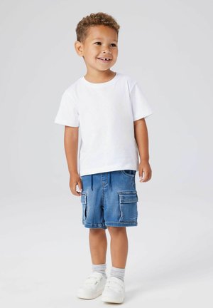 Jeune garçon souriant, debout, portant un t-shirt blanc, un short cargo bleu, des chaussettes grises et des baskets blanches sur un fond clair uni.