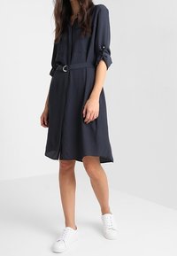 Abito camicia blu navy con chiusura a bottoni, maniche arrotolate e cintura in vita. Realizzato in tessuto leggero, abbinato a sneakers bianche.