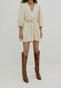Vestido crema envolvente con mangas abullonadas atado en la cintura, combinado con botas de tacón altas de imitación de cocodrilo en color marrón. Textura de tela suave.