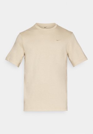 T-shirt in cotone beige chiaro con colletto rotondo, maniche corte e un piccolo logo Nike nero sul lato sinistro del petto. Texture liscia.