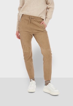 Un pantalon à cordon de serrage beige en matière douce, au design fuselé, doté de poches latérales et d'une texture lisse, associé à des baskets blanches.