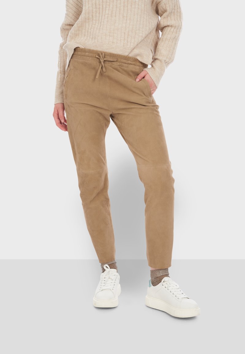 Tan trekbroek van zacht materiaal, met een taps toelopend ontwerp, zijzakken en een gladde textuur, gecombineerd met witte sneakers.