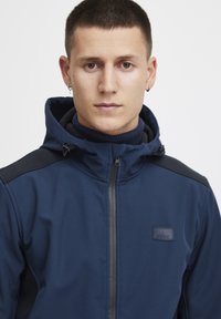 Tumši zila zip-up jaka ar augstu apkakli, melniem plecu akcentiem un mazu logo uzlīmi uz krūtīm. Gluda tekstūra.