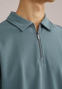 Svetlo modra, teksturirana pulover s zadrgo. Ima zapiranje s polovično zadrgo in oblikovan ovratnik. Narejen iz zračnega materiala.