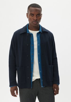 Navy blue cardigan with a collared neckline, button closure, and two front pockets, layered over a denim shirt with visible stitching.