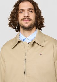 Bežna jakna z zadrgo in ovratnikom, na sprednjem delu z manjšo barvno oznako na levi strani prsnega koša. Notranjost prikazuje ovratnik s plavim karo vzorcem.