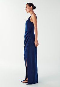 Moteris su be rankovių tamsiai mėlynu sukniu su šonine iškirpte, stovinti profiliu prieš pliką šviesią foną, plaukai surišti žemai į kuodą.