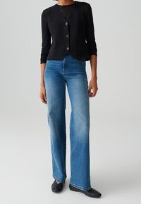 Černý bezrukávový veston s předními knoflíky, kombinovaný se světle modrými širokými džíny. Textury vypadají hladce s minimálním poškozením.