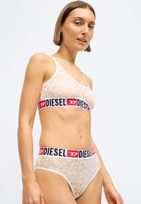 Δαντελένιο bralette και αντίστοιχο σλιπ σε κρέμα, με φαρδιά μαύρη και κόκκινη ζώνη με την επωνυμία "DIESEL". Το ύφασμα είναι ημι-transparent με λουλουδάτα σχέδια.