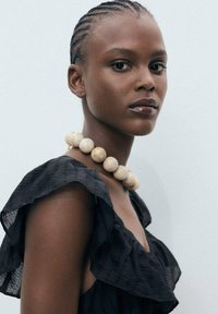Collier de perles beiges, robe noire texturée avec des manches volantes, peau lisse, cheveux tressés et un fond neutre.