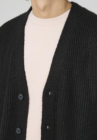 Gilet en tricot noir à côtes avec six boutons, porté sur une chemise à manches longues rose pâle. Le tissu texturé et le design à ouverture frontale sont visibles.