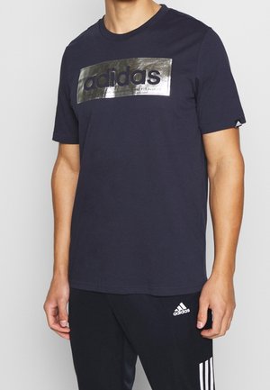 Navy-blaue Baumwoll-T-Shirt mit einem großen silbernen Adidas-Logo auf der Vorderseite, rundem Ausschnitt und kurzen Ärmeln. Kombiniert mit passenden dunklen Hosen.